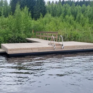 T‑mallinen putkiponttonilaituri mökkijärven rannalla; harmaa kestopuu ja uimaportaat.