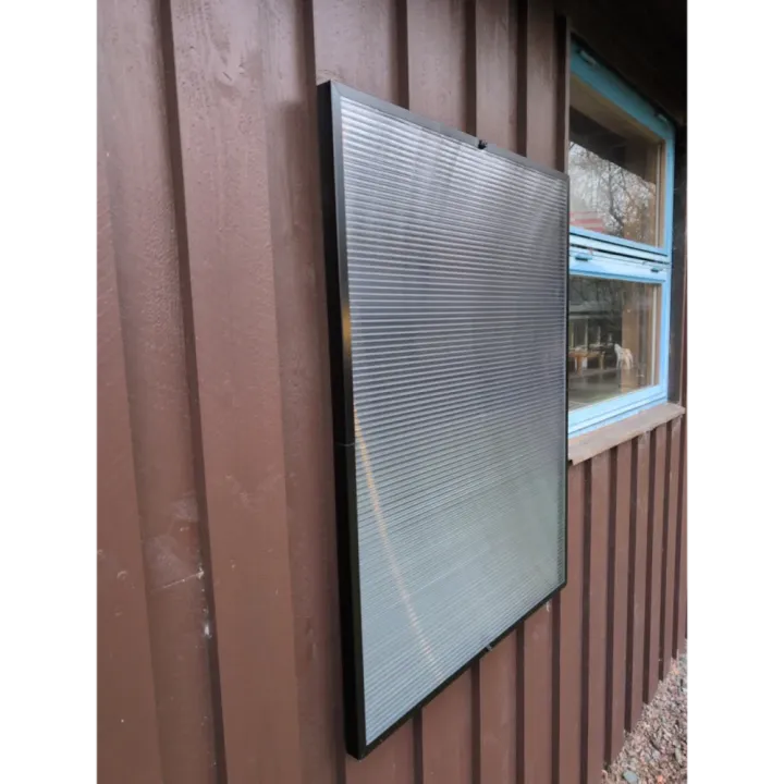 Täysmusta aurinkoilmakeräin asennettuna hirsiseinälle, valkoinen ilmaputki vie lämmintä ilmaa sisälle.